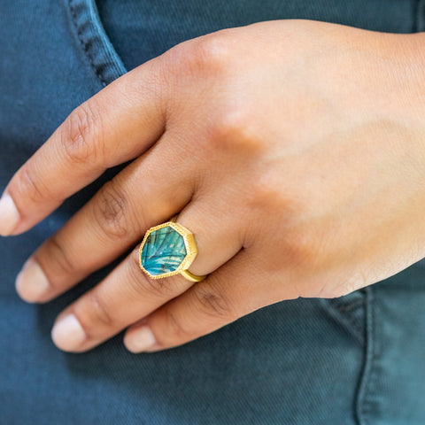   A model wears a labradorite ring hand-carved with an ornate pattern. The gemstone's gradient wash from blue to green shimmers in the light. Handmade 18K yellow gold bezel with braided detail. 