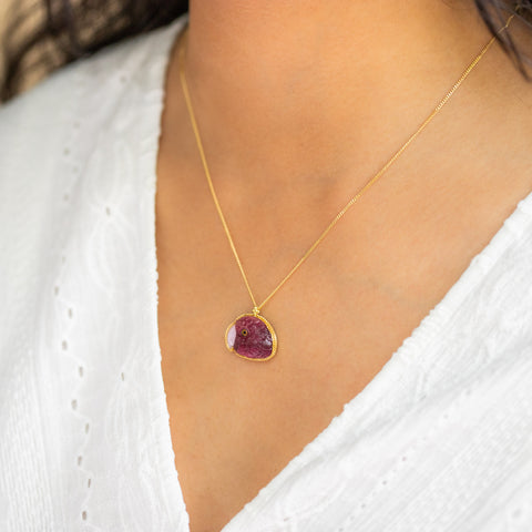 A model wears a captivating tourmaline parrot pendant in 18K yellow gold, hand-carved from magenta pink stone with detailed feathers, eyes, and Pink Opal beak. Suspended from a handmade gold bezel with braided detail on an 18K yellow gold chain. Handmade in New York