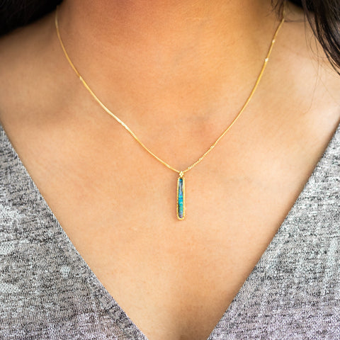 A model wears an Elongated Boulder Opal pendant which is set in an 18k yellow gold bezel, and suspended from an 18K yellow gold chain. Opal displays enchanting blues, purples, and greens, set in a unique one-of-a-kind design with braided detail.