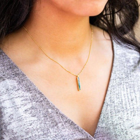 A model wears an Elongated Boulder Opal pendant which is set in an 18k yellow gold bezel, and suspended from an 18K yellow gold chain. Opal displays enchanting blues, purples, and greens, set in a unique one-of-a-kind design with braided detail.