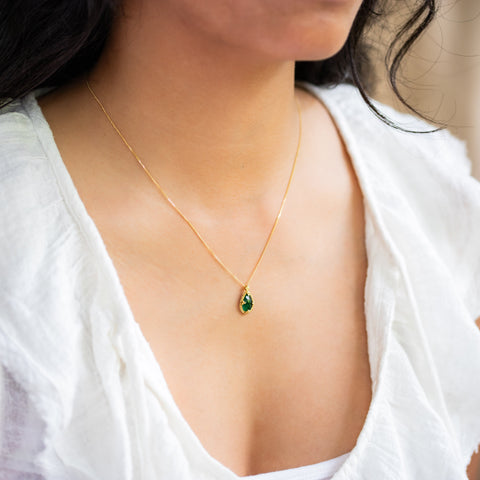 A model wears an emerald pendant with hues ranging from dark pine green to bright grassy green, reminiscent of a lush forest. Encased in an intricate frame of shimmering golden chain and delicate beaded prongs. Suspended on an 18k yellow gold chain, showcasing meticulous craftsmanship. Handmade in New York.