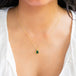 A model wears an emerald pendant with hues ranging from dark pine green to bright grassy green, reminiscent of a lush forest. Encased in an intricate frame of shimmering golden chain and delicate beaded prongs. Suspended on an 18k yellow gold chain, showcasing meticulous craftsmanship. Handmade in New York.