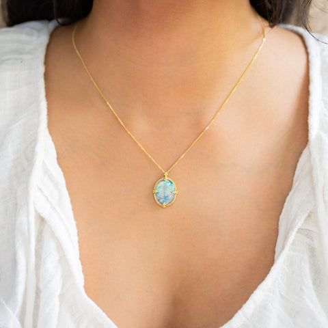 A model wears an Ethiopian Opal pendant in 18K yellow gold, revealing delicate iridescence and rainbow colors from electric green to peacock blue and neon-pink confetti. Suspended from a handmade gold frame with braided gold accents on an 18K yellow gold chain. Handmade in New York.