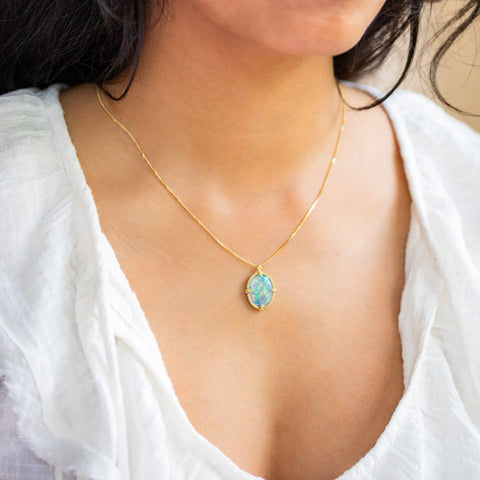 A model wears this Ethiopian Opal pendant in 18K yellow gold, boasting delicate iridescence. Rainbow colors from electric green to peacock blue and neon-pink confetti flicker across the surface. Suspended from a handmade gold frame with braided gold accents on an 18K yellow gold chain. Handmade in New York.