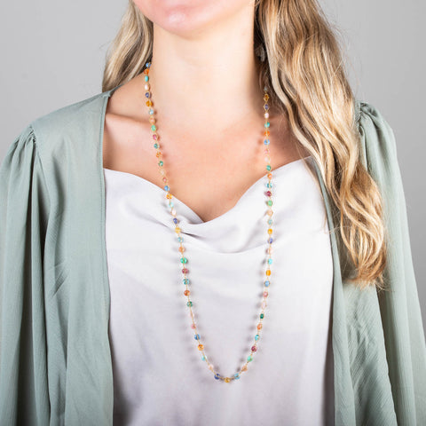 A model wears a multi-colored woven gemstone bead necklaces that features multiple gemstones including Aquamarine, Opal, apatite, Moonstone, Tanzanite, Carnelian, Tourmaline, Amazonite, Rhodochrosite, Chalcedony and Jade. The stones are set in a long 18k yellow gold chain.