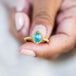 A model holds this Aquamarine ring in 18K yellow gold, highlighting a misty blue oval gemstone reminiscent of a tranquil summer sky. Handmade gold bezel with intricate braided gold accents and granulated prongs.