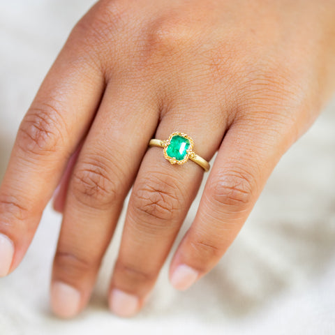 A model wears this Emerald ring in 18K yellow gold, capturing lush, vibrant green reminiscent of Spring sprouts. Intricate frame of shimmering golden chain and delicate beaded prongs. Handmade in New York.