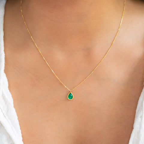 A model wears an emerald pendant with a glowing, perfect emerald green hue, suspended from an 18K yellow gold chain and set in a handmade gold bezel. Handmade in New York.