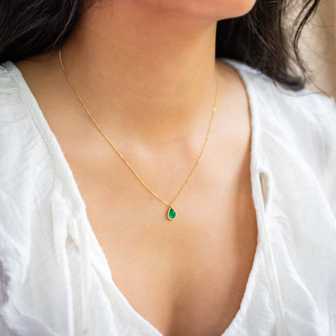 A model wears an emerald pendant with a glowing, perfect emerald green hue, suspended from an 18K yellow gold chain and set in a handmade gold bezel. Handmade in New York.
