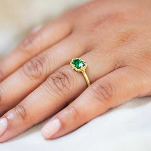 A model wears this Emerald ring in 18K yellow gold, its tiny facets reflecting light akin to dewdrops on green grass. Intricate frame of shimmering golden chain and delicate beaded prongs. Handmade in New York. 