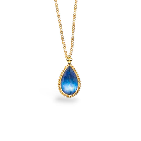 An iridescent teardrop-shaped Moonstone pendant encased in a handmade gold bezel with braided detail, capturing its captivating swirling blue beauty. Suspended on an 18k yellow gold chain, showcasing meticulous craftsmanship. Handmade in New York.