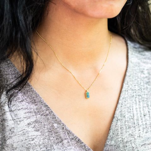 A model wears a rectangular Boulder Opal pendant set in 18k yellow gold bezel, suspended from an 18K yellow gold chain. Opal displays mesmerizing neon greens and blues, set in a unique one-of-a-kind design with braided detail.