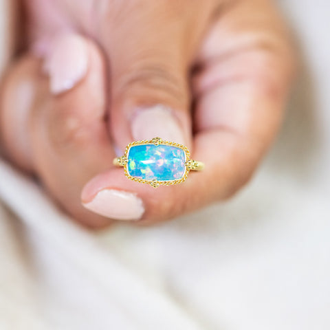 A model holds this Ethiopian Opal ring in 18K yellow gold, showcasing an organically shaped rectangular opal with a dazzling play of colors. Handmade gold frame features braided detail and granulations. Handmade in New York