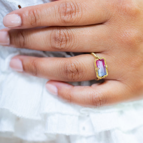 A model wears a Tourmaline ring in 18K yellow gold, revealing a captivating color shift from lavender to raspberry. Handmade gold frame displays delicate braided detail and granulation.