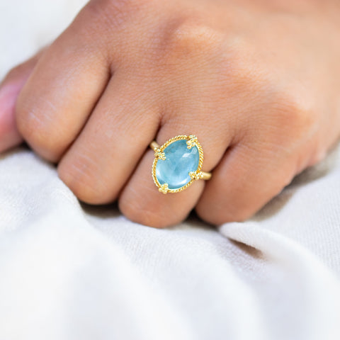 A model wears an aquamarine ring in 18K yellow gold, capturing the play of shades beneath the surface in the light. Handmade gold frame with braided gold accents and granulated prongs. Handmade in New York.