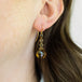 A close-up of an 18k yellow gold drop earring that features three woven black diamond beads and a teardrop shaped cognac quartz and hangs from a french hook closure