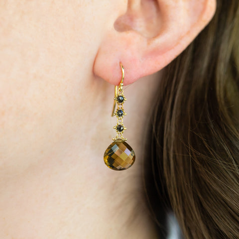 A model wears an 18k yellow gold drop earring that features three woven black diamond beads and a teardrop shaped cognac quartz and hangs from a french hook closure