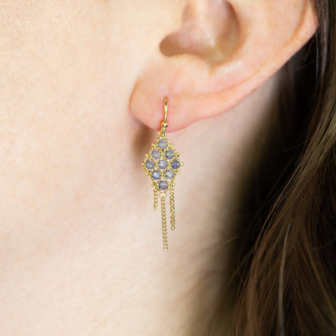 A model wears an 18k yellow gold earring crafted with tanzanite beads woven into a diamond shaped lattice pattern and has three dangling chains at the bottom.