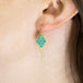 A model wears an 18k yellow gold earring crafted with turquoise beads woven into a diamond shaped lattice pattern and has three dangling chains at the bottom.