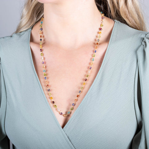 A model wears a long multi-colored stone necklace that is woven with 18k yellow gold chains. The necklace features gemstones including Sunstone, Peridot, Aquamarine, Lolite, Apatite, Imperial Topaz, Rhodolite Garnet, Morganite, Tanzanite, Amethyst, and Grossular Garnet