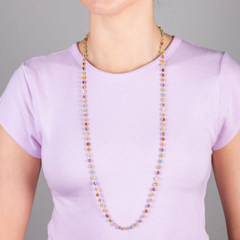 A model wears a 34" multi-colored woven gemstone necklace woven with delicate 18k yellow gold chains. The necklace features Sunstone, Peridot, Aquamarine, Lolite, Apatite, Imperial Topaz, Rhodolite Garnet, Morganite, Tanzanite, Amethyst, and Grossular Garnet