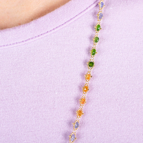 A close up of a long woven chain necklace that features Green Tourmaline, Garnet, and Tanzanite stone beads woven in 18k yellow gold chain.