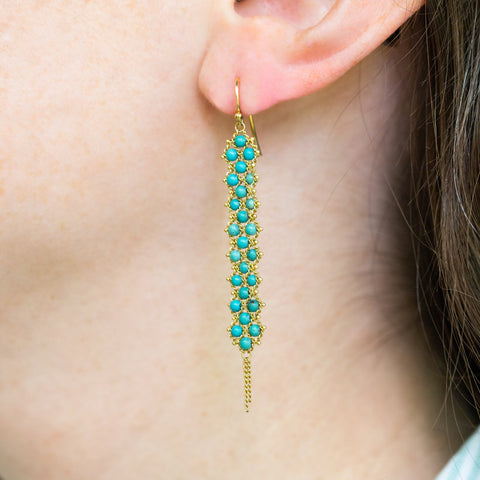 A model wears a pair of long turquoise earrings which feature a woven lattice pattern and two dangling chains at the bottom of the earring. The earrings fasten with french hook closures.