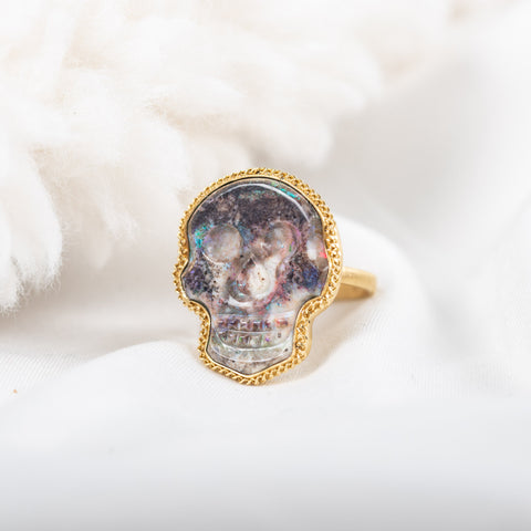 A large purple hued opal is carved into a skull and set in a braided 18k yellow gold bezel. The stone rests on a thin ring band.