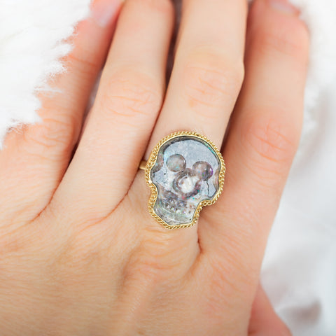 A model wears a large skull carved purple opal ring. The stone is set in a chain wrapped 18k yellow gold bezel and rests on a thin band.