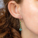 A model wears a drop earring featuring a row of emeralds woven together in 18k yellow gold with a teardrop shaped apatite stone at the bottom.