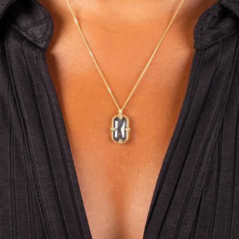 A vertical rectangular black diamond pendant is set in an 18k yellow gold chain wrapped bezel with four beaded prongs. The pendant is hanging on a delicate chain.