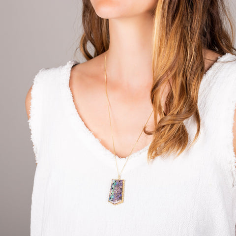 A model wears a large carved Andamooka opal, with dark purple, green and blue hues, that has a delicate scalloped edge motif. The pendant is set in an 18k yellow gold chain wrapped bezel and hangs on a delicate long chain.