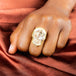 A model wears a large skull carved pink opal ring. The stone is set in a chain wrapped 18k yellow gold bezel and rests on a thin band.