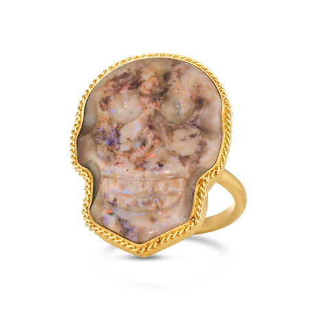 A large pink hued opal is carved into a skull and set in a braided 18k yellow gold bezel. The stone rests on a thin ring band.