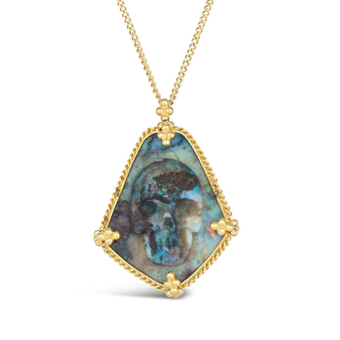 An andamooka opal, with blue and black hues, has a skull carved into the stone. The pendant is set in an 18k yellow gold chain wrapped bezel with four beaded prongs and hangs on a delicate short chain.