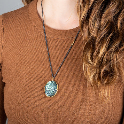 A large labradorite stone is carved with a floral design and set in an 18k yellow gold bezel. The pendant hangs from an oxidized sterling silver chain with three off center blue diamonds set in the chain.