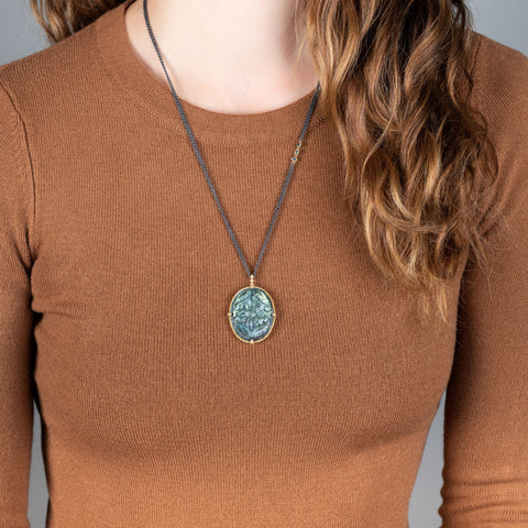 A model wears a large floral carved labradorite stone set in 18k yellow gold and hanging from an oxidized sterling silver chain with three off center blue diamonds.
