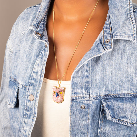 A model wears a carved owl pendant made from light pink rhodochrosite with a blue beak and gold and black eyes on a long delicate chain.