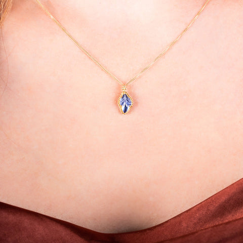 A small tanzanite stone carved like a leaf is set in an 18k yellow gold bezel with a braided detail around the edge and hangs from a delicate chain.