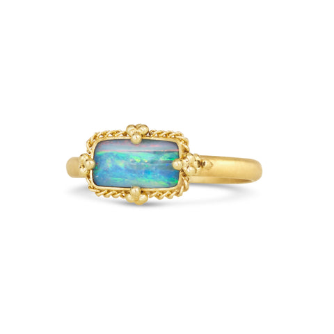 A rectangular opal stone with blue, green and peach streaks is set in an 18k yellow gold chain wrapped bezel with four beaded prongs on a thin band.