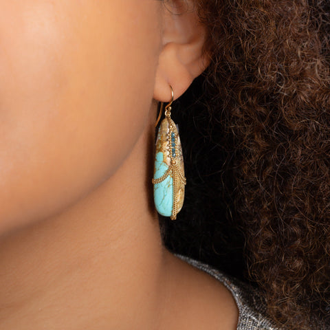 A model wears a large turquoise teardrop shaped earring. The stone is wrapped in an 18k yellow gold chain with blue diamonds set in the chain.