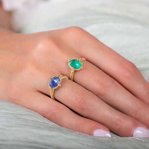 A model wears a small oval shaped emerald ring set in an 18k yellow gold chain wrapped bezel. The stone sits on a thin band.