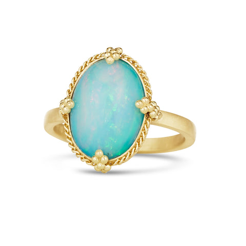 A large oval shaped Ethiopian opal with blue hues is set in an 18k yellow gold chain wrapped bezel with four beaded prongs and sits on a thin ring band.