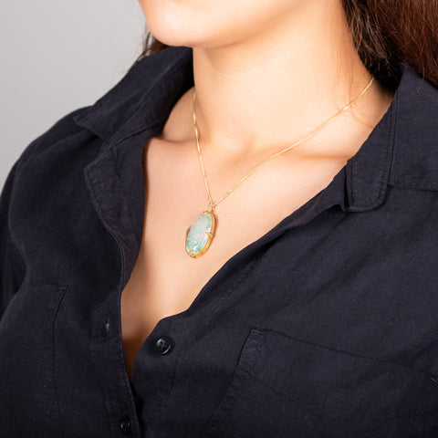 A model wears a large oval shaped Ethiopian opal pendant that is set in an 18k yellow gold chain wrapped bezel with four beaded prongs. The pendant hangs on a delicate short chain.
