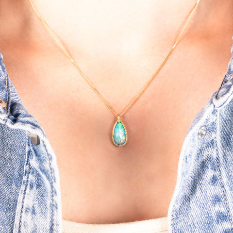 A teardrop shaped Ethiopian opal pendant is set in an 18k yellow gold chain wrapped bezel with four beaded prongs. The pendant hangs on a delicate chain.