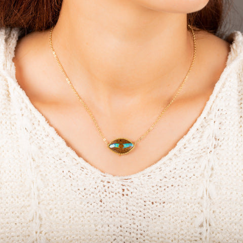 A model wears a medium eye shaped opalized wood pendant, featuring a blue streak through the center, and is set in 18k yellow gold chain wrapped bezel. The pendant is suspended on a delicate chain. 