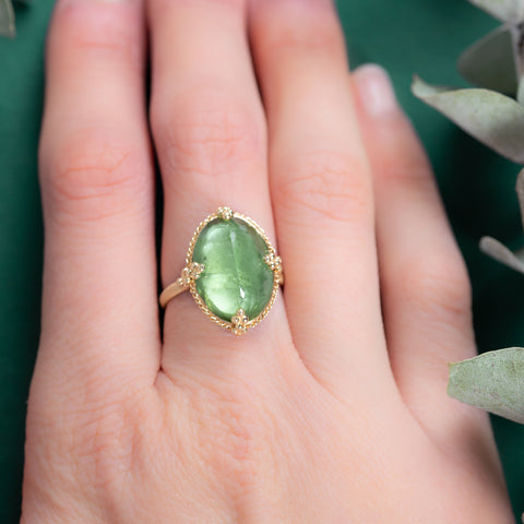 A model wears a large oval shaped green tourmaline ring is crafted in 18k yellow gold and set in a chain wrapped bezel with four beaded prongs on a thin band.