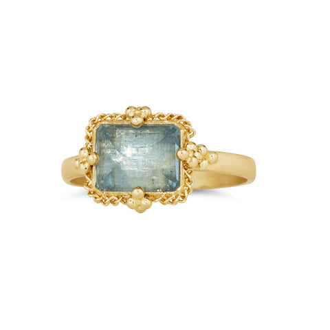 A rectangular faceted aquamarine ring is set in an 18k yellow gold chain wrapped bezel with four beaded prongs that sits on a thin band.