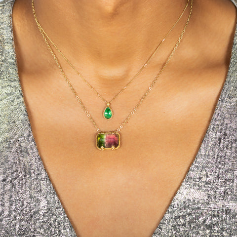 A model wears two layered necklaces, including a teardrop shaped emerald pendant and a rectangular watermelon tourmaline necklace.