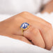 A model wears a large tanzanite stone ring is set in an 18k yellow gold chain wrapped bezel with four beaded prongs. The stone sits on a thin band.